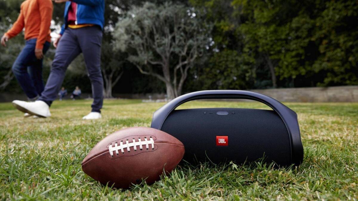 Портативная колонка Harman JBL на зеленой траве с футбольным мячом сбоку и игроками на заднем плане