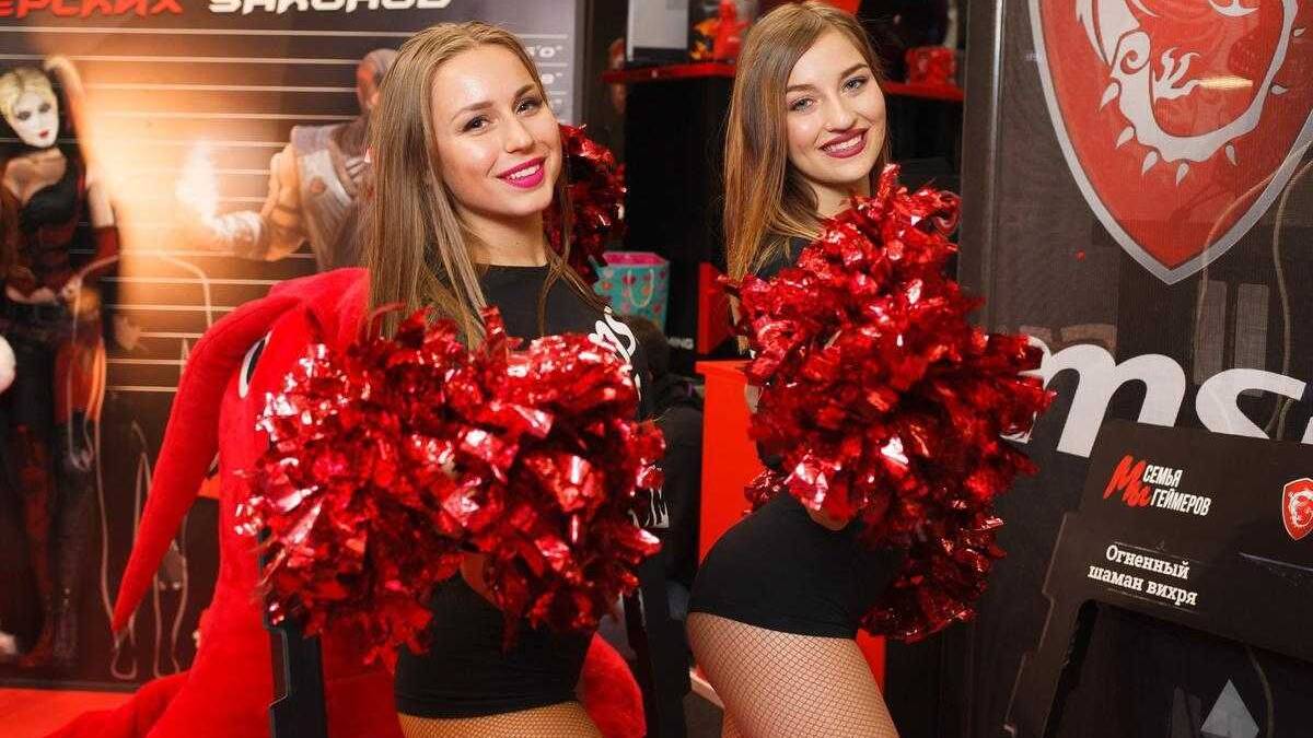 18+ Самые красивые девушки ИгроМир 2017 и Comic Con 58 Две улыбающиеся девушки в чёрных костюмах с красными украшениями и помпонами на фоне логотипа и выставочного стенда