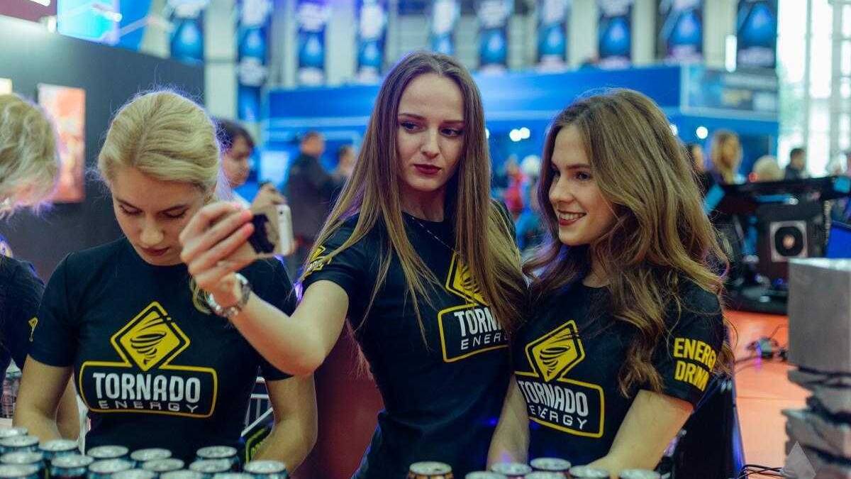 18+ Самые красивые девушки ИгроМир 2017 и Comic Con 78 Трое девушек в черных футболках с логотипом Tornado Energy, одна делает селфи у стойки с энергетическими напитками