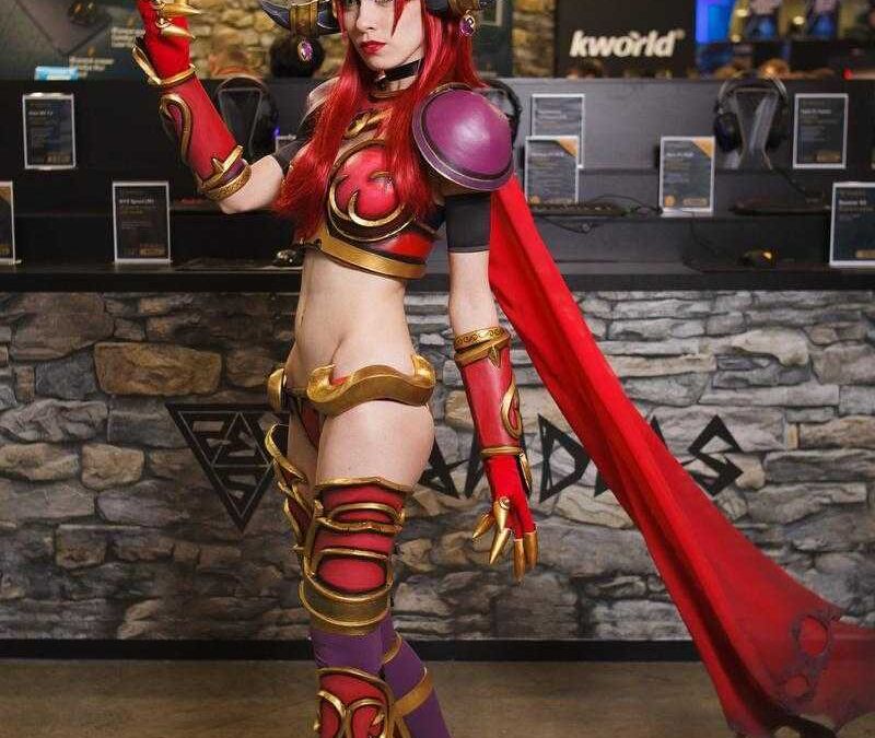 Косплей персонажа из игры с фантазийным костюмом и шлемом рогов, стоящий у стойки игровой выставки