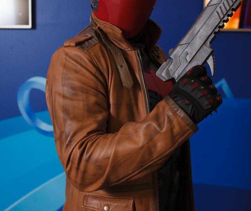 Человек в красной маске и кожаном пиджаке держит пистолет на фоне синего стены