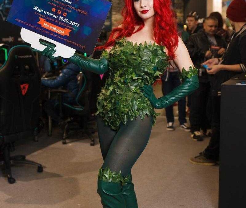 Косплей героини из игровой параллели с красными волосами и зелеными аксессуарами на выступлении