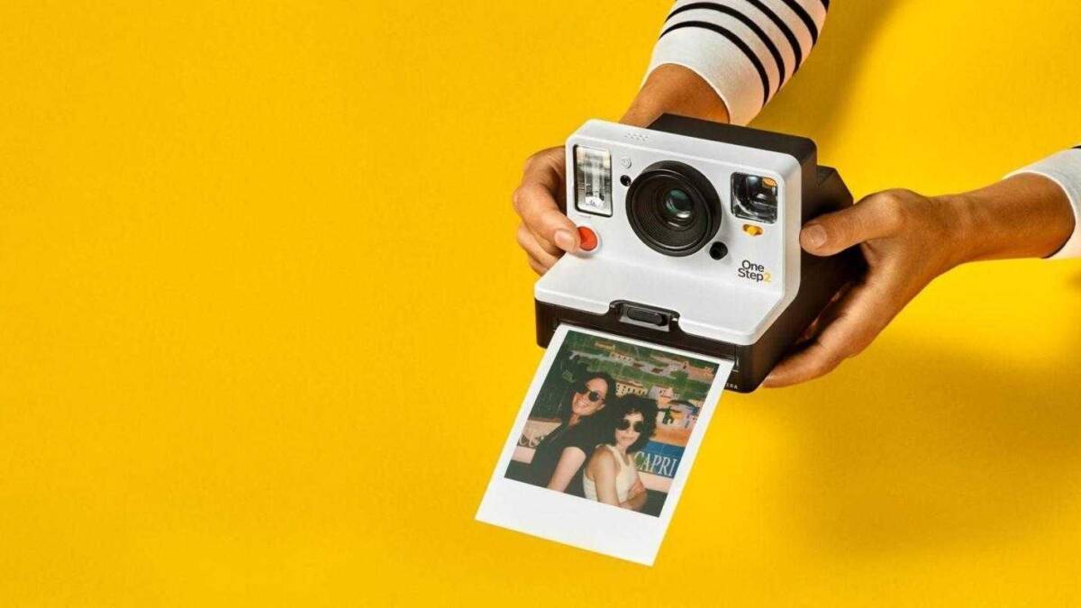 Человек держит фотокамеру Polaroid OneStep+ с напечатанным фото на ярком желтом фоне