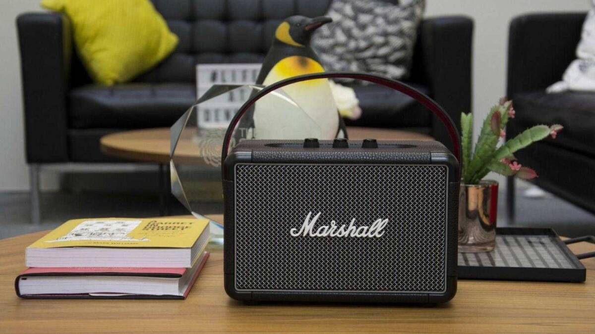На столе стоит портативная акустика Marshall Kilburn II со стальным решетчатым фронтальным покрытием и логотипом Marshall