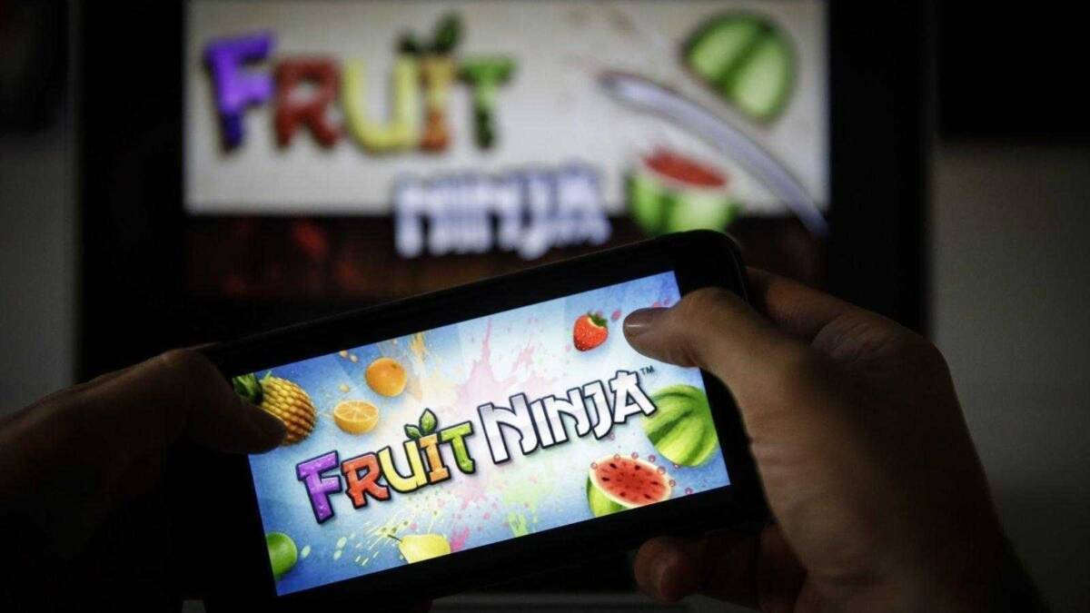 Человек держит смартфон с логотипом Fruit Ninja и изображениями фруктов на экране