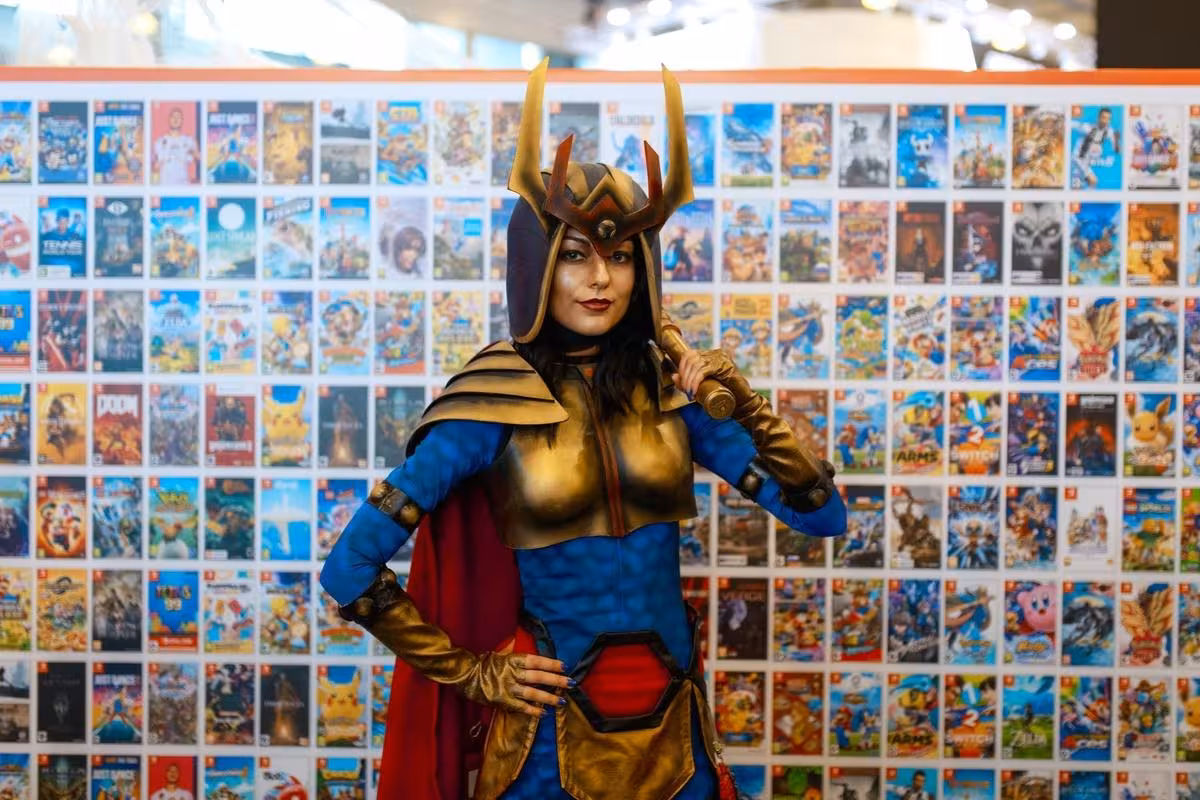 Косплей персонажа с короной в костюме синего и красного цветов на фоне плакатов игр