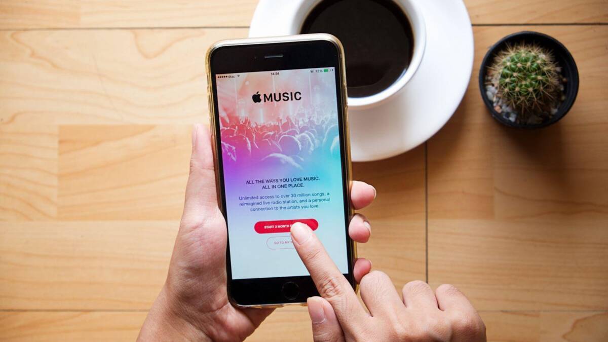 Рука держит смартфон с приложением Apple Music на экране, на столе возле чашки кофе и кактуса