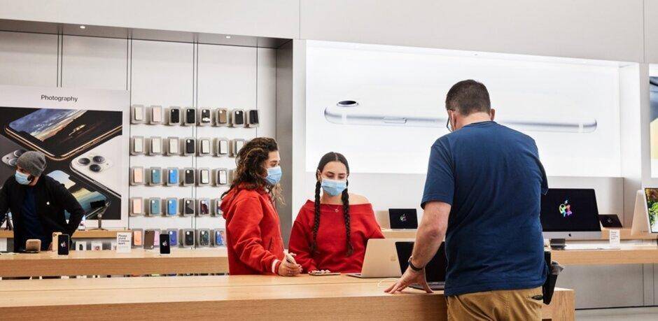 Apple открывает 25 магазинов Apple Store в США 3 Мужчина и две женщины в масках в магазине Apple Store за прилавком с ноутбуками и iPhone