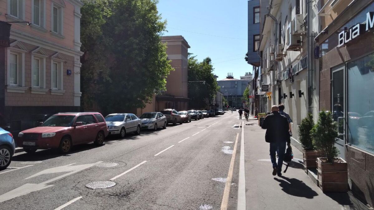 Улица с припаркованными автомобилями и пешеходами рядом с магазинами iPhone 12