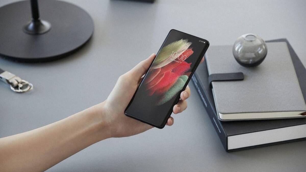 Флагманские смартфоны Galaxy S21 поступили в продажу в России 4 Рука держит смартфон Samsung Galaxy S21 Ultra на фоне настольных книг и аксессуаров