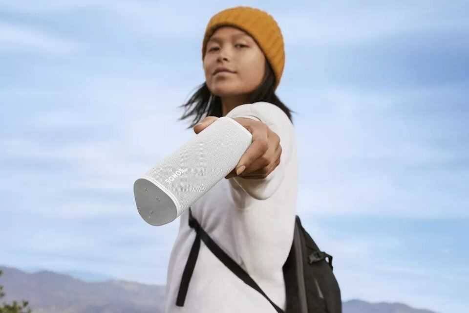 Девушка с желтым шапкой держит портативную колонку Sonos Roam впереди