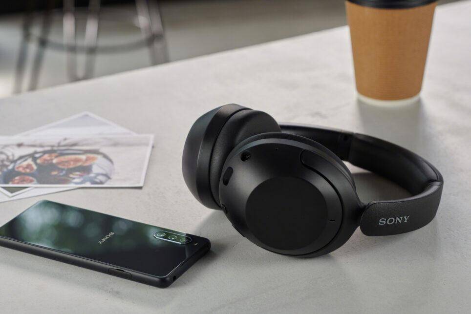 Черные наушники Sony WH-XB910N на столе рядом с мобильным телефоном и фотографиями