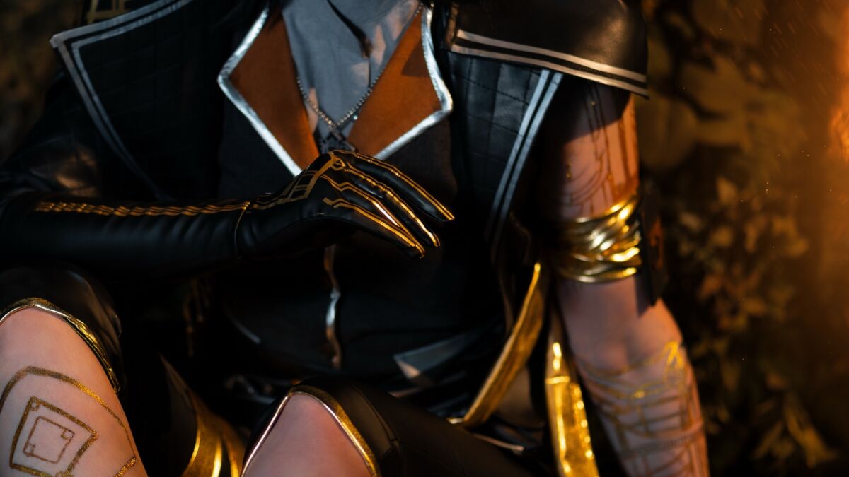 Косплей Люнэ из Clair Obscur Expedition 33 Lizzie Whistle девушка с черными волосами в костюме с золотыми акцентами и татуировками на ногах на тёмном фоне