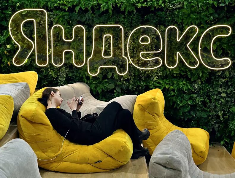 Женщина лежит на желтом диване в кафе с зеленой стеной и неоновым знаком Яндекс