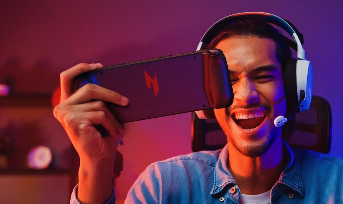 Молодой человек держит смартфон Acer Nitro Blaze Link с игровой клавиатурой и мышью у ноутбука