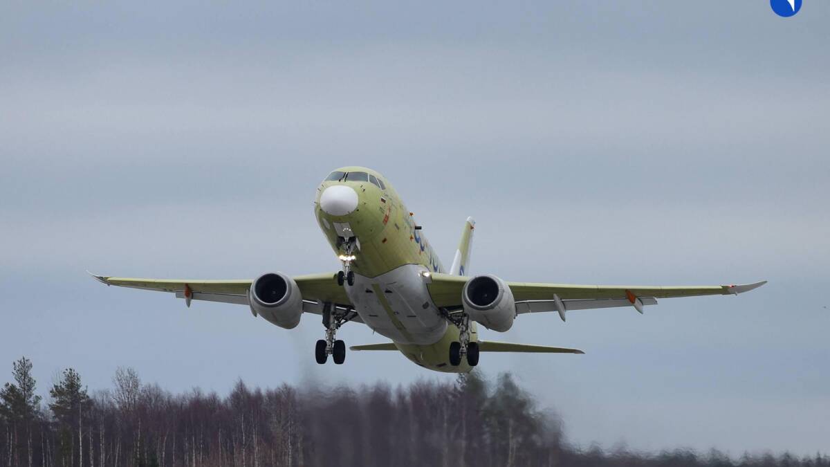 Самолет SJ-100 в полёте над деревьями на фоне облачного неба